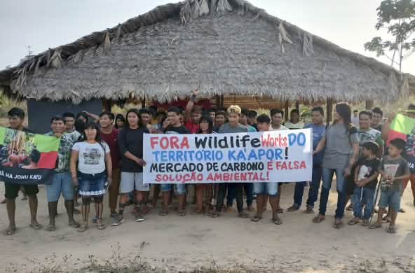 Más de 30 indígenas Ka'apor de pie frente a una casa tradicional con pilares de madera y techo de paja. Las personas sostienen una pancarta de protesta en la que se lee: "¡Fuera Wildlife Works del territorio ka'apor! El mercado de carbono es una falsa sol