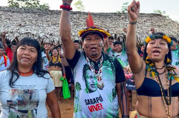 Tres indígenas Ka'apor —dos mujeres y un hombre, con los puños en alto delante de un espacio cubierto con hojas de palma.