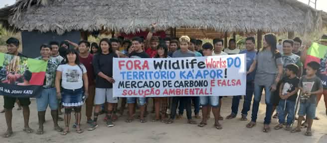 Más de 30 indígenas Ka'apor de pie frente a una casa tradicional con pilares y techo de paja. Las personas sostienen una pancarta en la que se lee: "¡Fuera Wildlife Works del territorio ka'apor! El mercado de carbono es una falsa solución ambiental”