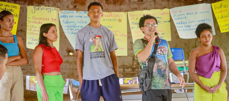 Jóvenes de la Amazonia del estado brasileño de Maranhão participan en talleres de comunicación para defender sus territorios Cinco jóvenes, tres mujeres y dos hombres, de pie, frente a un tendedero en el que cuelgan carteles. Uno de los jóvenes habla por un micrófono que sujeta con la mano derecha.