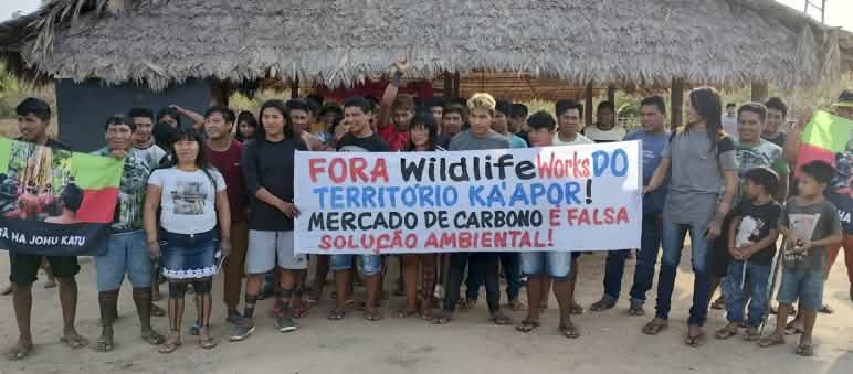 Manifestación de indígenas Ka'apor contra la presencia de la empresa Wildlife Works en el territorio Más de 30 indígenas Ka'apor de pie frente a una casa tradicional con pilares y techo de paja. Las personas sostienen una pancarta en la que se lee: "¡Fuera Wildlife Works del territorio ka'apor! El mercado de carbono es una falsa solución ambiental”